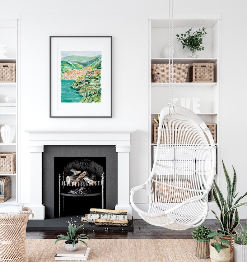 May include: A framed watercolor painting of a coastal town with green hills and turquoise water hangs above a fireplace. A white wicker hanging chair and built-in shelves with baskets and plants complete the cozy, bright interior.