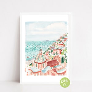 May include: Watercolor print of a coastal town with buildings cascading down a hillside to the turquoise sea. The artwork features a domed building and a light blue sky. The print is framed in a white border.