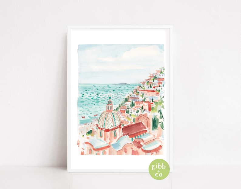 May include: Watercolor print of a coastal town with buildings cascading down a hillside to the turquoise sea. The artwork features a domed building and a light blue sky. The print is framed in a white border.