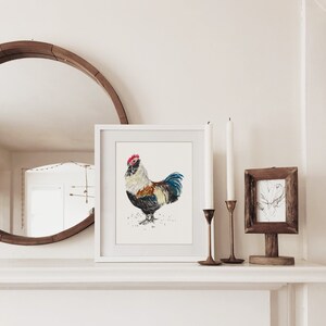 May include: A white framed print of a watercolor painting of a rooster with a red comb and black, white, and brown feathers. The rooster is standing on a white background.