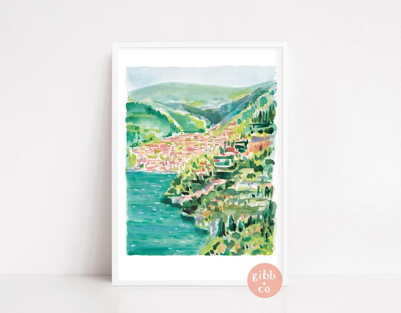 May include: Watercolor painting of a coastal town with red-roofed buildings nestled along a turquoise body of water. Lush green hills and trees surround the town. The artwork is framed in a white border.