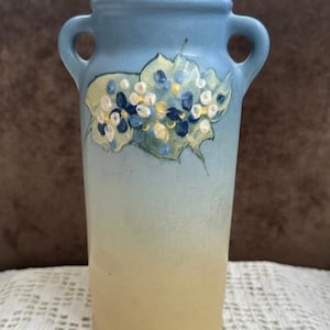 May include: A ceramic vase with a light blue and beige gradient. The vase features two handles and is decorated with a painted floral design in shades of blue, white, and yellow. The vase is approximately 8 inches tall.