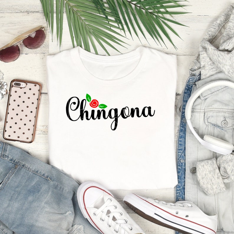 Op de afbeelding: Wit T-shirt met de tekst "Chingona" in zwarte cursieve letters en een rode roos met groene bladeren boven de tekst.