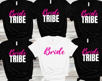 May include: Five black t-shirts with the text "Bride Tribe" in white and one white t-shirt with the text "Bride" in pink. The shirts are arranged in a semi-circle on a wooden surface.