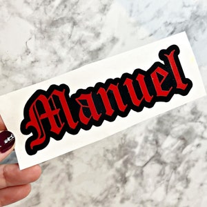 May include: A red vinyl sticker with the name "Manuel" in black outlined gothic lettering.