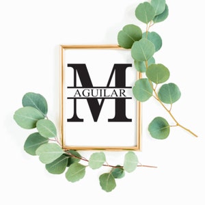 May include: A gold framed print with a black letter M and the word AGUILAR centered below the letter. The print is on a white background and is surrounded by green eucalyptus branches.