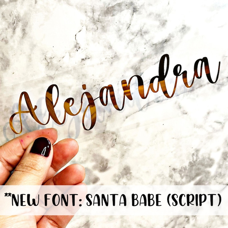 May include: Gold vinyl sticker with the name "Alejandra" written in a cursive font called "Santa Babe (Script)".