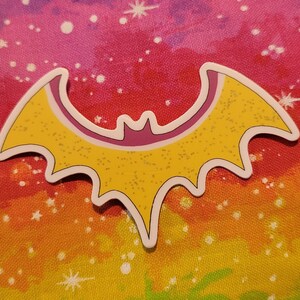 May include: A yellow bat-shaped sticker with a white outline and glitter accents. The sticker has a purple interior and the text "passion fruit" in a yellow rectangle. The background is a colorful rainbow pattern.