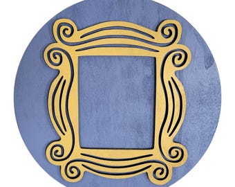 May include: A round, blue wooden wall hanging with a gold-colored frame in the center. The frame has a square opening and decorative swirls. A natural rope is attached for hanging.