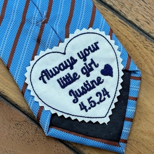 May include: A white heart-shaped fabric patch with blue embroidery that reads "Always your little girl Justine 4.5.24" attached to a blue and brown striped tie.