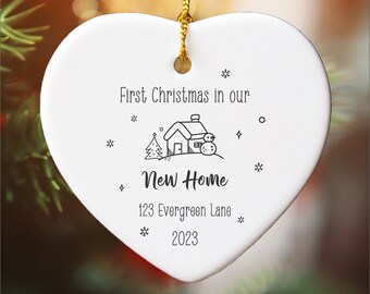 May include: White heart-shaped ceramic ornament with black text that reads "First Christmas in our New Home 123 Evergreen Lane 2023". A small black and white illustration of a house with a tree and snowman is above the text.
