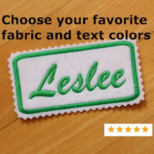 Może przedstawiać: Zielona haftowana etykieta z imieniem z białego materiału z imieniem "Leslee" w zielonym, ozdobnym pismem.