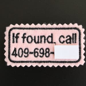 Könnte beinhalten: Ein rosa Filzanhänger mit schwarzem Rand und gezackten Kanten. Der Anhänger lautet "If found, call 409-698-" mit einem leeren weißen Rechteck darunter. Der Text ist fett und ohne Serifen.