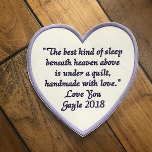 May include: A white heart-shaped patch with purple stitching. The patch has a quote embroidered on it: "The best kind of sleep beneath heaven above is under a quilt, handmade with love." The patch also has the text "Love You Gayle 2018" embroidered on it.