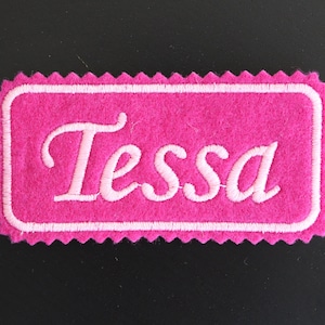 May include: Pink felt name tag with white stitching, featuring the name "Tessa" in a cursive font.