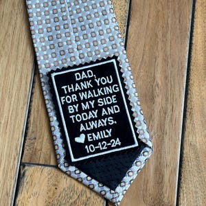 May include: A black tie with a white and brown patterned design. A black patch with white text reads "DAD, THANK YOU FOR WALKING BY MY SIDE TODAY AND ALWAYS. EMILY 10-12-24".