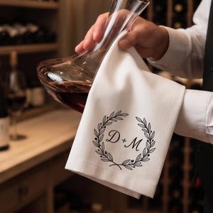 May include: A white cloth napkin featuring a black printed laurel wreath design surrounding the letters D + M. The napkin is draped over a clear glass wine decanter containing a dark red liquid, held by a person wearing a white shirt and waistcoat.