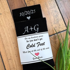 May include: Black socks with white embroidered text and a decorative label. The socks display the date "10/20/25", initials "A+G", and the phrase "So you don't get Cold Feet". A small heart graphic is also present.