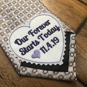 May include: A white heart-shaped fabric patch with the text "Our Forever Starts Today 11.4.19" embroidered in purple thread. The patch is attached to a brown and white patterned tie.