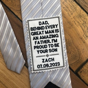 May include: A gray and white striped tie with a white patch that reads "Dad, Behind Every Great Man Is An Amazing Father. I'm Proud To Be Your Son Zach 07.09.2023".