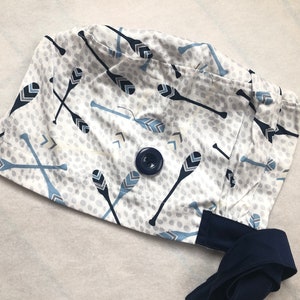 May include: A white scrub cap featuring a pattern of navy and light blue canoe paddles and arrows. The cap has a navy blue button and a navy blue tie. The fabric has a dotted background.