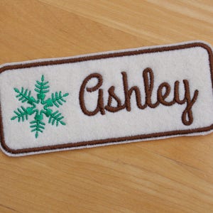 Pode incluir: Um patch retangular branco com uma borda marrom e o nome "Ashley" bordado em itálico marrom. Um desenho de floco de neve verde é bordado no lado esquerdo do patch. O patch é provavelmente para uma jaqueta ou bolsa.