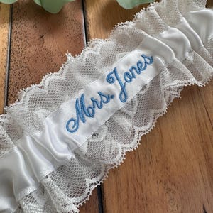 May include: White satin and lace wedding garter with the embroidered text "Mrs. Jones" in light blue script. The garter features a delicate lace trim and a satin band, perfect for a bride.