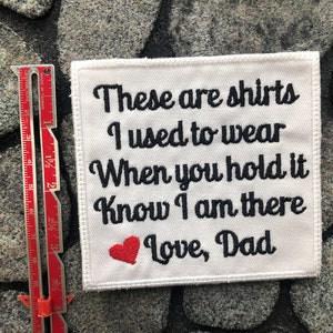 Op de afbeelding: Witte geborduurde patch met zwarte tekst die "These are shirts I used to wear When you hold it Know I am there Love, Dad" luidt. Een rood hart is onder de tekst geborduurd.