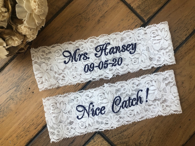 A set of pretty white lace garters embroidered with the bride's new last name and wedding date.  On the top keepsake garter , the embroidery says Mrs. Hansey and below that is the wedding date.  The throw garter says "Nice Catch".