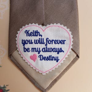 May include: A beige tie with a white heart-shaped patch sewn on. The patch has pink stitching around the edges and says "Keith, you will forever be my always Destiny" in blue embroidery.