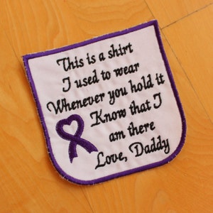 Op de afbeelding: Witte stoffen patch met paarse stiksels en een paars lint met een hartvorm. De patch heeft de tekst "This is a shirt I used to wear Whenever you hold it Know that I am there Love, Daddy."