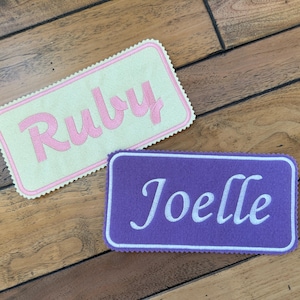 Peut inclure: Deux étiquettes de nom personnalisées, l'une avec le nom "Ruby" en lettres roses sur un fond jaune clair et l'autre avec le nom "Joelle" en lettres blanches sur un fond violet.
