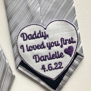Könnte beinhalten: Eine silber gestreifte Krawatte mit einem herzförmigen Aufnäher. Der Aufnäher zeigt einen lila gestickten Text: "Daddy, I loved you first. Danielle 4.6.22" zusammen mit einem lila Herzen.