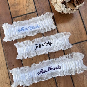 May include: Three white lace garters with gathered fabric centres. The top garter features blue embroidery reading Mrs Clarke, the middle garter has black embroidery reading Jeff's Wife, and the bottom garter has purple embroidery reading Mrs. Favela.