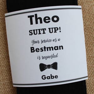 May include: A white card with black text that reads "Theo Suit Up! Your service as a Bestman is requested Gabe" with a black bow tie graphic.