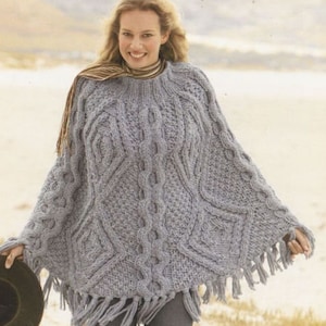 Op de afbeelding: Een grijze gebreide poncho met een kabelbreipatroon en franjes. De poncho wordt gedragen door een persoon met lang bruin haar.