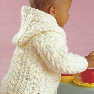 May include: A cream-coloured, hooded baby sweater with a cable knit design. The sweater features a hood and long sleeves. The baby is wearing khaki trousers. The background is a light pink colour.