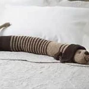 Puede incluir: Una almohadilla térmica de punto con forma de perro de rayas marrones y beige. La almohadilla está sobre una colcha blanca.