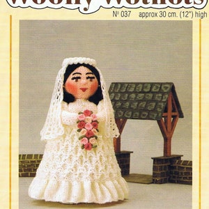 Puede incluir: Una muñeca de novia de crochet blanco con velo, sosteniendo un ramo de flores rosa y blanco. La muñeca mide 30 cm de alto y está de pie frente a una pequeña estructura de madera marrón.