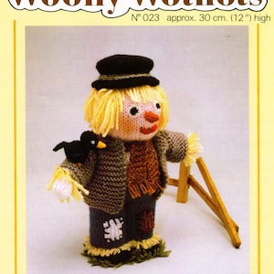 May include: A knitted scarecrow doll with a black hat, yellow hair, and a brown scarf. The doll is holding a small black bird and is standing next to a wooden fence. The doll is 12 inches tall.