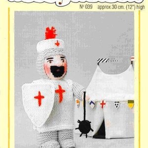 Puede incluir: Patrón de tejido para un muñeco cruzado blanco y gris con un escudo de cruz roja, sosteniendo una bola negra con púas. El patrón se titula "Woolly Wotnots" y es el número 039. El muñeco mide aproximadamente 30 cm de alto.