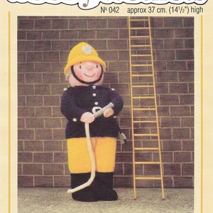 Może przedstawiać: Dzianinowa figurka strażaka, o wysokości około 37 cm, w żółtym hełmie, granatowej kurtce, żółtych spodniach i czarnych butach, trzymająca biały wąż. Żółta drabina stoi obok figurki. Widoczny tekst "Woolly Wotnots" i "THE FIREMAN".