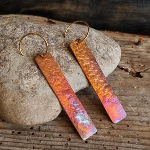 Op de afbeelding: Een paar rechthoekige oorbellen met een gehamerde textuur en een kleurovergang van oranje, roze en paars. De oorbellen hebben goudkleurige oorhaken. De oorbellen worden gepresenteerd op een stenen oppervlak.