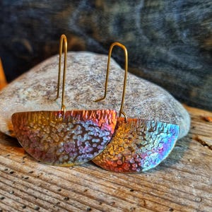 Puede incluir: Pendientes artesanales con un acabado iridiscente único. Los pendientes tienen forma de media luna con una superficie texturizada, mostrando una mezcla de colores dorados, rojos y azules. Los ganchos para las orejas son de metal dorado simple.