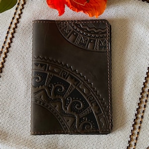 May include: A brown leather passport holder with an intricate embossed design. The holder is lying on a white and brown fabric with a floral design.