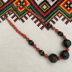May include: A necklace featuring coral-colored beads, black carved beads, and dark metal accents. The necklace is displayed against a backdrop of a white fabric with a colorful embroidered pattern.