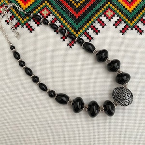 May include: A black beaded necklace with a silver pendant featuring a swirling design. The necklace is on a white fabric background with a colorful embroidered pattern.