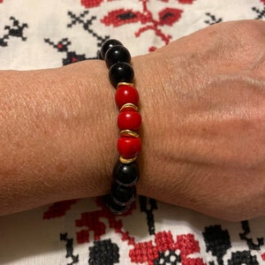 May include: A black and red beaded bracelet with gold accents. The bracelet is made of round beads and is worn on a person's wrist.