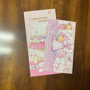 May include: Two pink and white greeting cards with illustrations of a baby in a crib and a baby in a pink blanket. The text on the cards reads "Z narodzhennyam donechky!" and "Z narodzhennyam donechky!"