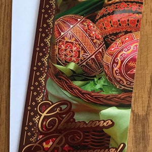 May include: Easter greeting card featuring intricately decorated eggs in a woven basket with green ribbon and paper grass. The card has a brown border with gold accents and a greeting in gold script. The card is on a white envelope.